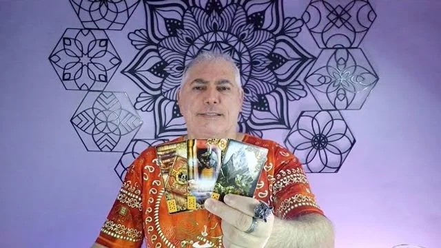 Henrique T’Oyá de camisa laranja estampada, com três cartas na mão, diante da parede de mandalas