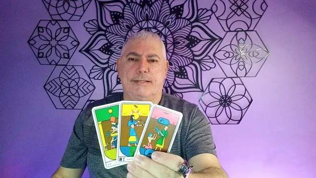 Henrique T’Oyá de camisa cinza, mostrando três cartas de tarô coloridas, diante da parede de mandalas