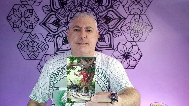Henrique T’Oyá de camisa branca, mostrando uma carta ilustrada com uma figura de vermelho sob uma árvore, diante da parede de mandalas