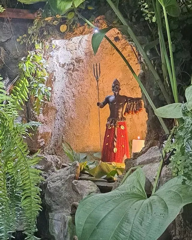 O jardim do Templo: uma gruta entre folhagens, com uma imagem de Exu e uma vela acesa